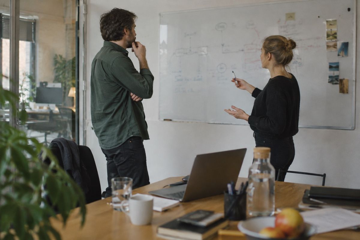 Symbolbild: Ein Mann und eine Frau stehen in einem Besprechungsraum vor einem Whiteboard mit skizzierten Abläufen, im Vordergrund ein Tisch mit aufgeklapptem Laptop