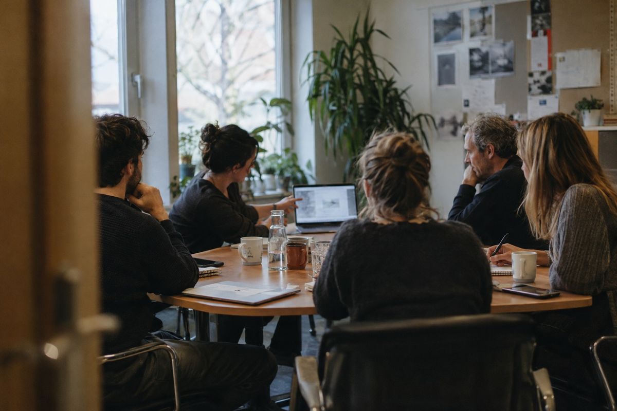 Symbolbild: Fünf Kolleginnen und Kollegen sitzen an einem runden Besprechungstisch, eine von ihnen zeigt etwas auf ihrem Laptop, die anderen hören zu und machen Notizen
